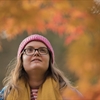 A woman stands in an Autumnal area, looking up at the red and orange leaves. 