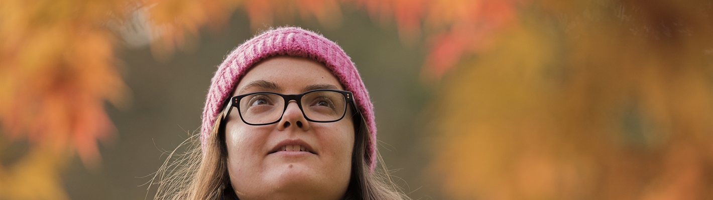 A woman stands in an Autumnal area, looking up at the red and orange leaves. 