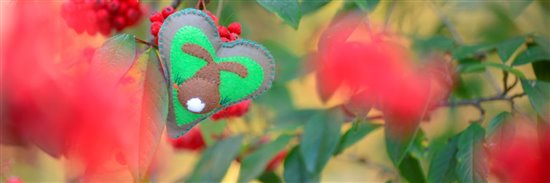 A grey padded heart, with a blue blanket stitch border, depicting the back of a rabbit (complete with white fluffy tail) surrounded by carrots on a green background. The heart is balanced amongst the branches of a bush full of bright red berries.