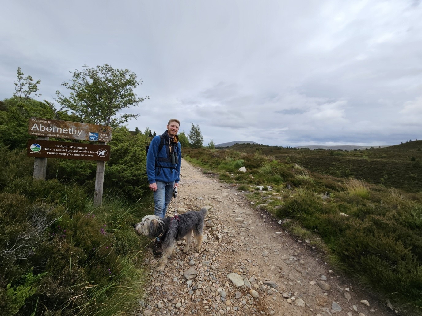 RSPB Scotland's Ben Oliver Jones is standing on a track with his dog. They are by a sign which reads, "Welcome to Abernethy National Nature Reserve".