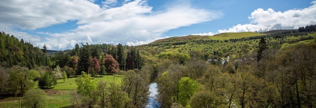 A glistening river flows through a lush green valley surrounded on either side by wooded slopes. Above is a bright blue sky with white fluffy clouds.
