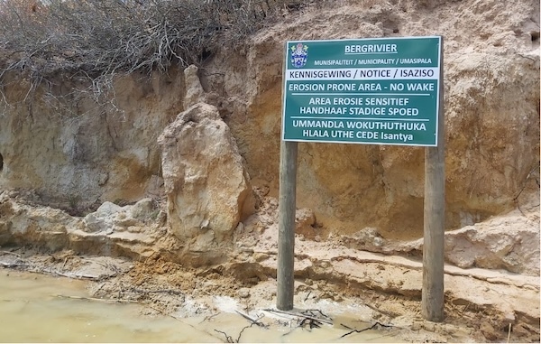 A sign saying 'Erosion Prone Area - No Wake' stands in front of an eroded riverbank