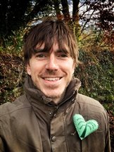 TV presenter Simon Reeve smiles at the camera. He is wearing a khaki coat with a handcrafted green heart pinned to his chest.