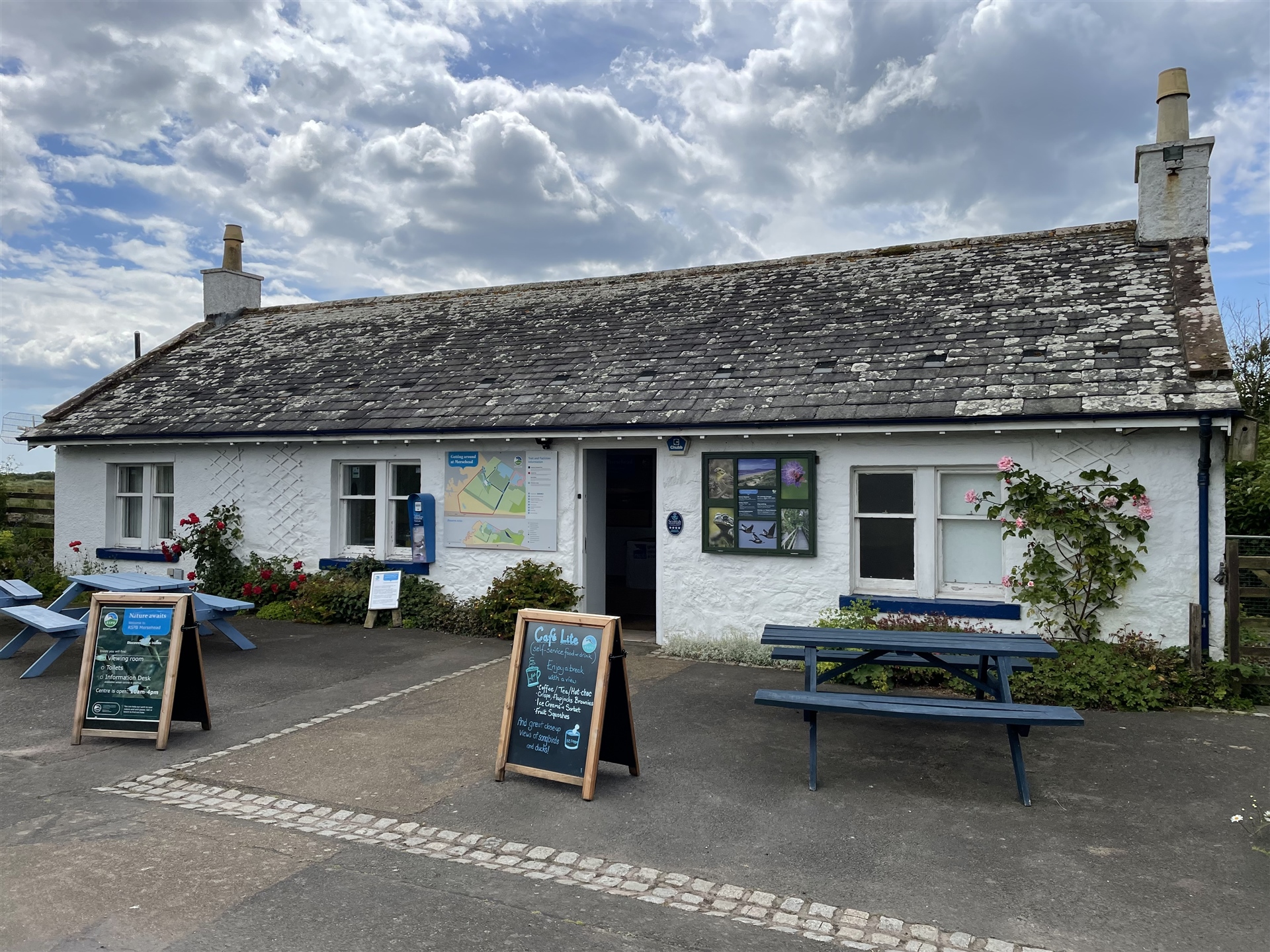 The visitor centre at RSPB Scotland Mersehead. It is a white cottage with maps, signs and benches outside.