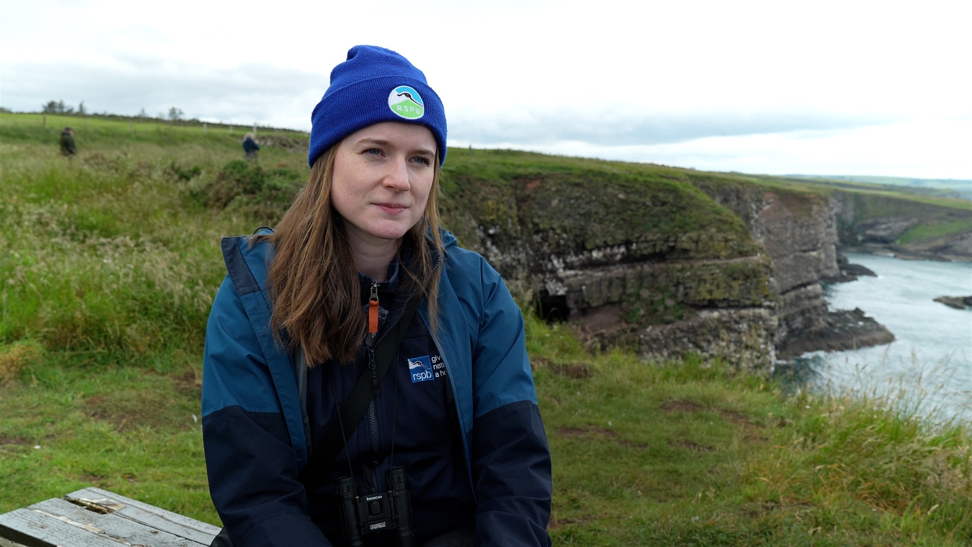 Caitlin looking off camera with cliffs in the background. She has an RSPB branded hat on.