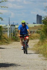 RSPB volounteer Rob is cycling along the Rainham Marshes River Wall, in the background there is a view across a section of the reserve and in the distance the buildings of London.