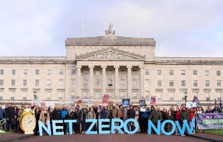A large group of people stand in front of the Stormont building, holding large letters that spell out Net Zero Now.