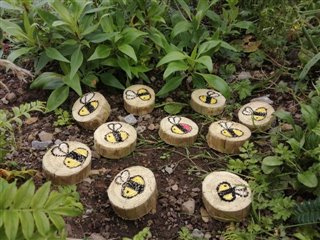 Ten little bees have been painted on to ten slices of wood and are placed on the ground with lush green plants all around them.