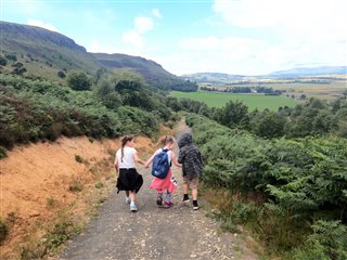 Three children holding hands walking down a path. The view behind them shows trees, hills and fields under a blue sky.