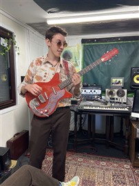 a person with short hair, a bright coloured t shirt, sunglasses, and brown trousers, holding a bright red guitar and standing in front of sound mixing equipment