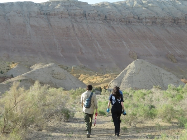 Two people walk towards a large outcrop of rock in a desert-like landscape