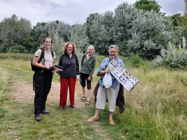 Four people smile at the camera. They are holding field guides and clipboards for a butterfly survey. They are in a grassy area with dense scrubby vegetation in the background.