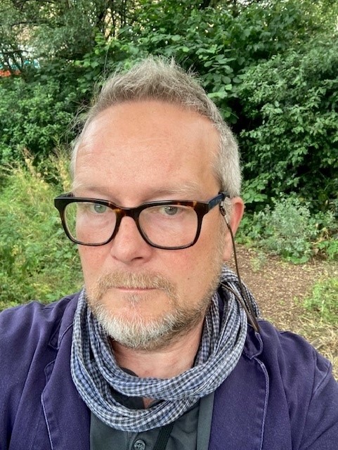 Selfie of Paul Walton, he is standing outside and wears glasses, a gingham nexk scraf, grey grandad t-shirt and blue linen jacket