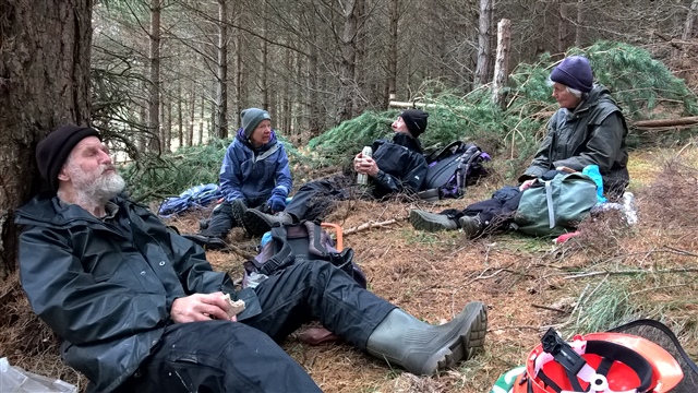 A group of four people in raincoats sit on the forest floor with flasks and sandwiches. Felled trees are in the background