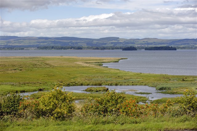 A panoramic view of RSPB Scotland Loch Leven, with wetlands and trees in the foreground, followed by the loch itself then hills in the distance.