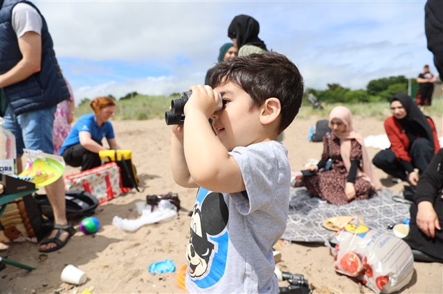 A young boy is looking through binoculars, while families are having picnics on the beach behind.