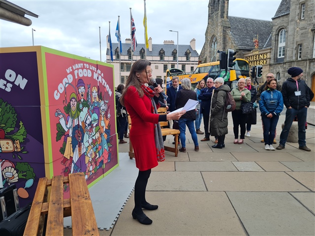 Arianne Burgess is speaking to a gathering of people outside the Scottish Parliament. She is stood next to a sign which reads, "We want to live in a good food nation".