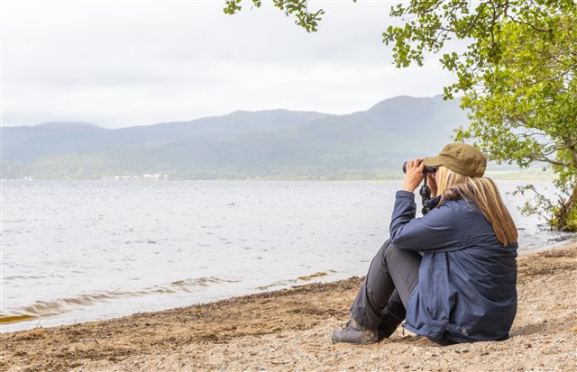 Volunteer with binoculars on beach at Loch Lomond. 