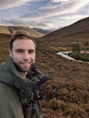 David smiling at the camera in front of a heather-covered landscape. He is wearing binoculars around his neck.