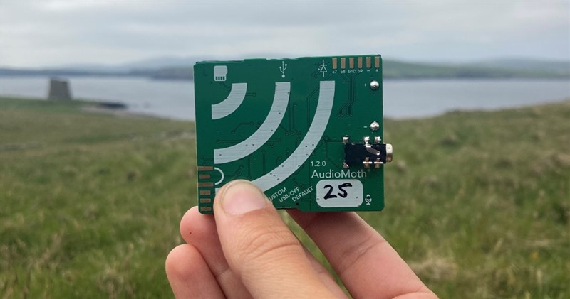 A hand holds up a green bit of board which has various electical components attached to it. Mousa broch can be seen in the background 