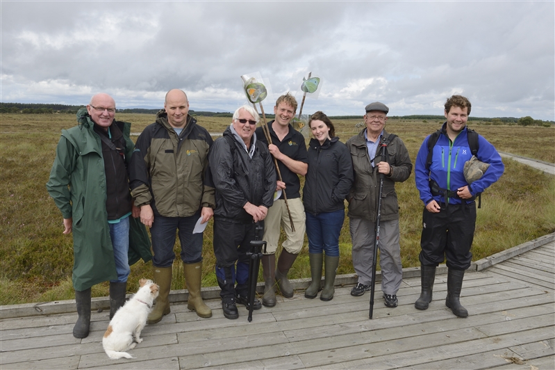 A group of people in outdoor clothing stand on a boardwalk above a peatland bog.